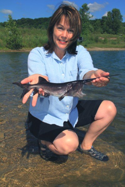 Woman holding a catfish while kneeling in shallow water, surrounded by greenery and a clear blue sky, showcasing a successful fishing experience.