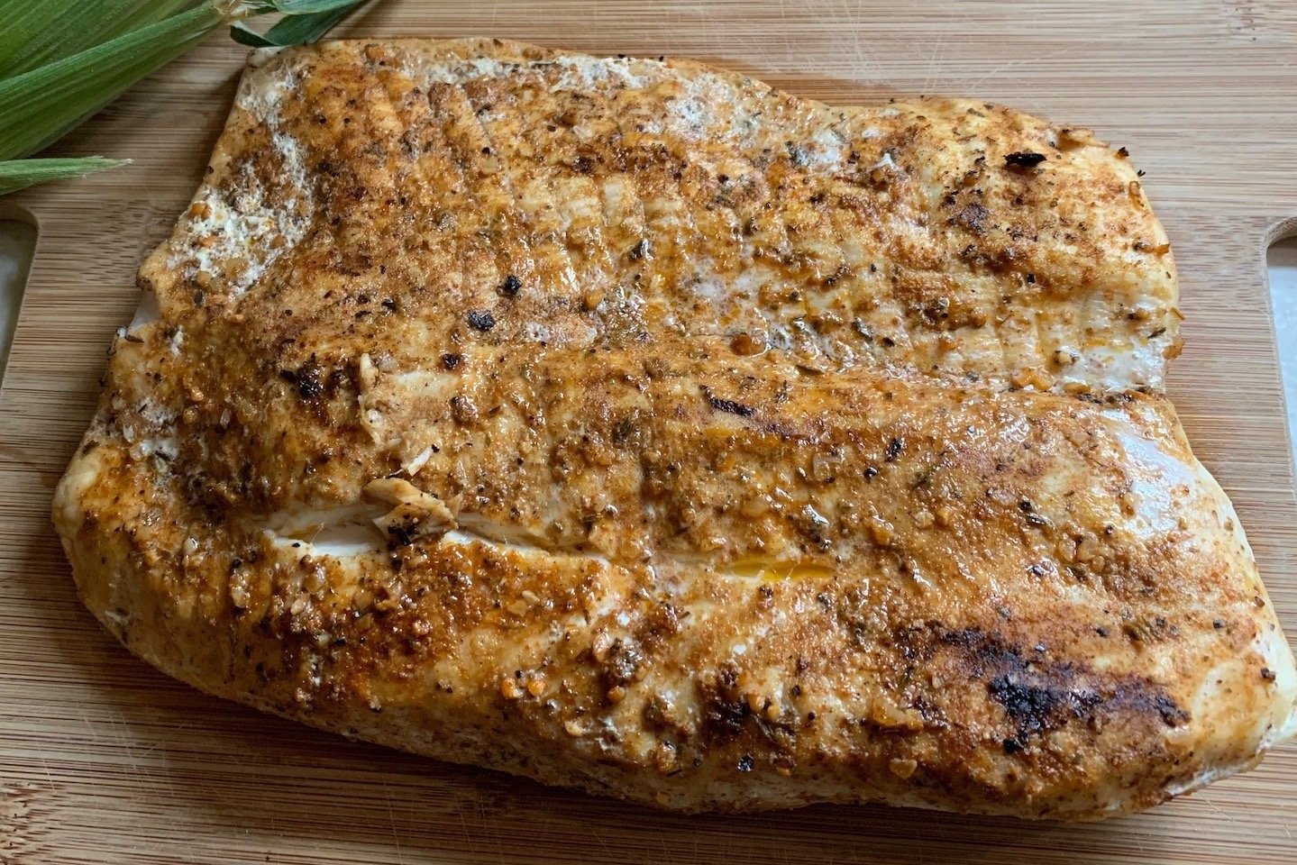 Grilled seasoned fish fillet on a wooden cutting board, showcasing a golden-brown crust and spices, with fresh green herbs in the background.